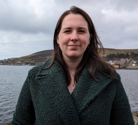 Woman in a green coat standing by a body of water with a backdrop of hills and buildings on a cloudy day.