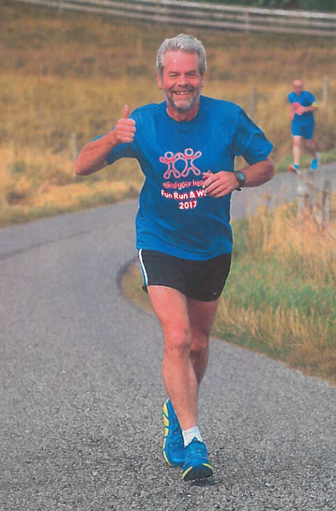 A man with a beard and mustache runs on a paved road, giving a thumbs-up and smiling. He is wearing a blue "Fun Run & Walk 2017" t-shirt, black shorts, and blue running shoes.