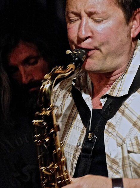 A man playing a saxophone with closed eyes, wearing a plaid shirt. Another person with long hair is in the background, partially visible.