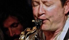 A man plays a saxophone with his eyes closed, while another person with long hair appears in the background.