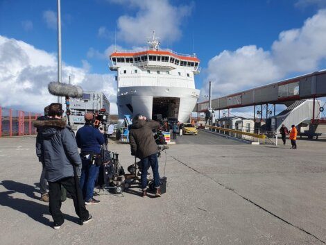 People filming in front of a large ferry docked at a port, with equipment and crew visible in the foreground.