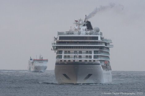 Two cruise ships are sailing on the sea, with one in the foreground and the other in the distance. Smoke is coming from the stack of the nearer vessel. The sea appears calm under a gray sky.