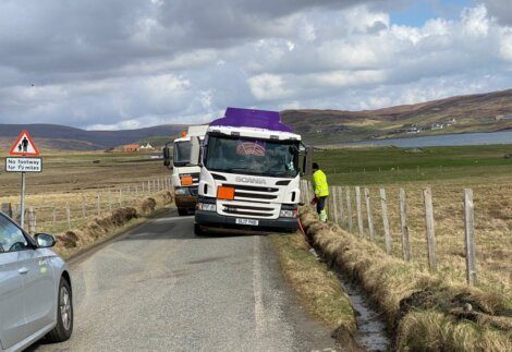 A truck with a purple roof is tilted off the road, with a person in a high-visibility jacket standing next to it. Another vehicle is on the same road, with a "No footway for 2 miles" sign visible.