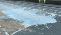 A large spill of white liquid, likely paint, covers a section of a road. The spill spans across the lanes, spreading unevenly. Nearby pavement and surroundings appear unaffected.