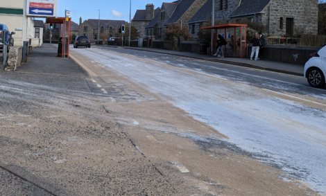 A road in a residential area is partially covered with sand or salt. Pedestrians are seen waiting at a bus stop on the right, and a car is approaching an intersection in the background.