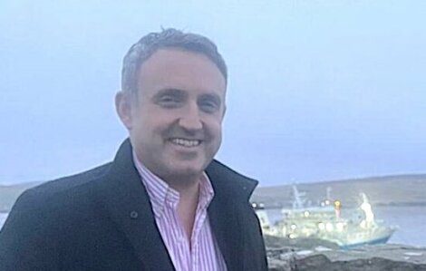 A person wearing a dark coat and pink shirt smiles in front of a blurred sea view with a boat in the background.