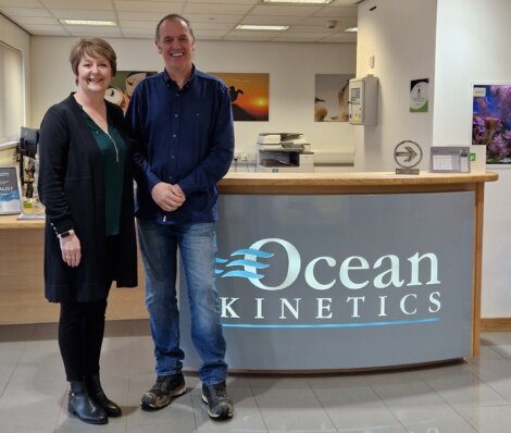 Two people standing and smiling in front of the reception desk of Ocean Kinetics.