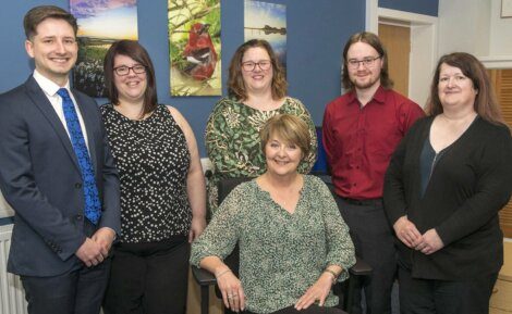 A group of five people stands behind a seated woman, all posing for a photo indoors. They are dressed in business casual attire, with framed pictures on the wall behind them.