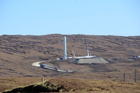 A wind turbine under construction in a hilly, barren landscape with construction equipment and cranes visible around the site.