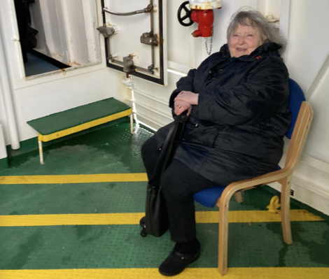 An older woman in a black coat sits on a chair indoors, beside a green and yellow-patterned floor and a red fire hydrant.