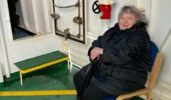 An elderly woman wearing a black coat sits on a chair in what appears to be a ship's interior. She is holding a folded black umbrella and smiling at the camera.