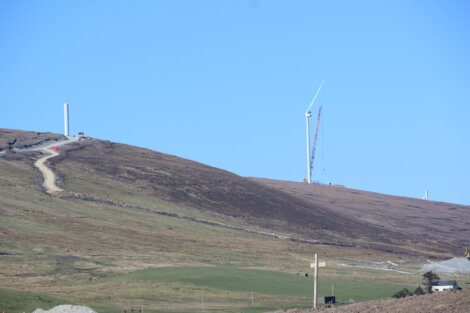 A construction site on a hill with a partially-assembled wind turbine, a winding path leading up, and clear blue sky above.