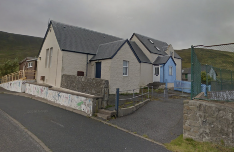 A small cluster of houses with white and blue exteriors stand along a paved road; a fence and a low wall are visible in the foreground.