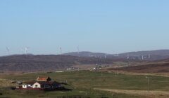 A rural landscape with a small house in the foreground and several wind turbines on rolling hills in the background.