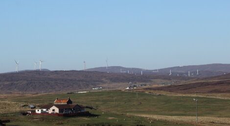 A rural landscape with a small house in the foreground and several wind turbines on rolling hills in the background.