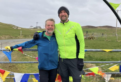 Two men, dressed in running gear, smile and stand outdoors near colorful bunting flags. A rural landscape with a house and wind turbine is visible in the background.