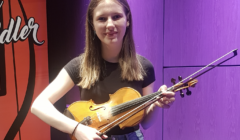 A young woman holding a violin, standing against a purple-lit backdrop. She is wearing a black t-shirt and jeans.