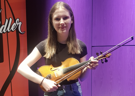 A young woman holding a violin, standing against a purple-lit backdrop. She is wearing a black t-shirt and jeans.