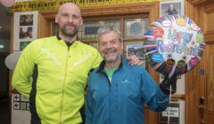 Two men stand in front of a "Happy Retirement" banner and hold a "Happy Retirement" balloon. Both are smiling; one wears a yellow jacket, the other a blue jacket.