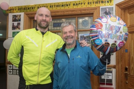 Two men stand in front of a "Happy Retirement" banner and hold a "Happy Retirement" balloon. Both are smiling; one wears a yellow jacket, the other a blue jacket.