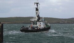 A small tugboat named "Einar" navigates through choppy waters near a coastal area with rolling hills in the background.