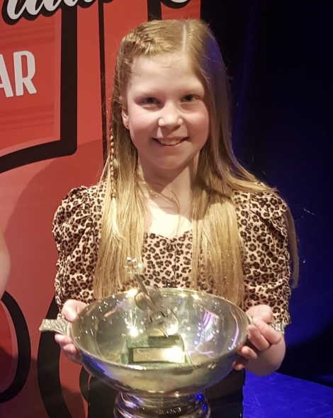 A young girl with long blonde hair and a leopard print top holds a large silver trophy, smiling at the camera.