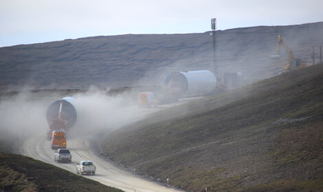 Construction site on a hilly road with heavy machinery, large cylindrical structures being transported by trucks, and a white vehicle driving through a dusty environment.