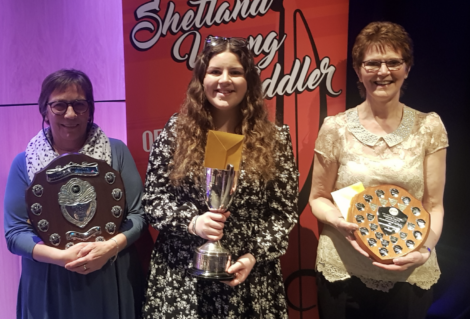 Three people stand holding awards; the middle person holds a trophy, and the others hold shield plaques. A sign behind them reads "Shetland Young Fiddler.