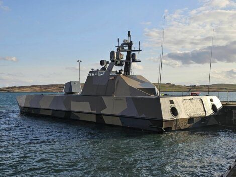 A modern military vessel, P865, with a camouflage pattern, docked at a harbor on a sunny day. The ship is equipped with antennas and various equipment on its deck.