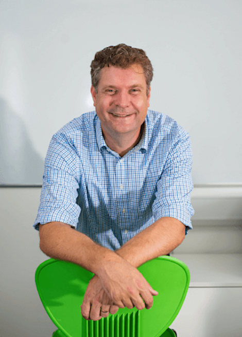 A man in a blue checkered shirt smiles while leaning on the back of a bright green chair.