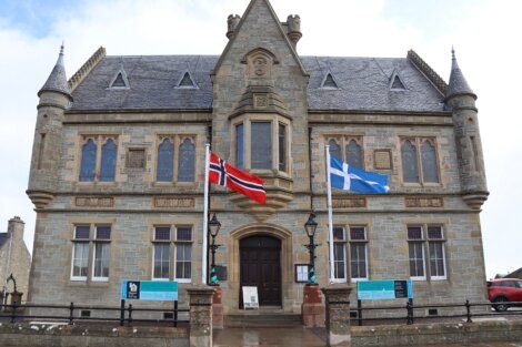 A historic stone building features two flags, Norway's on the left and Scotland's on the right, flanking the entrance. The structure has pointed arch windows and a symmetrical design.