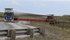 A truck transports a large wind turbine blade on a rural road, navigating a sharp turn with the blade extending over a fenced field.