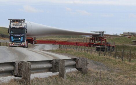 A truck transports a large wind turbine blade on a rural road, navigating a sharp turn with the blade extending over a fenced field.