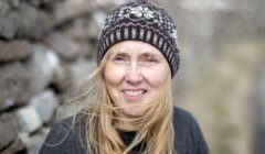 A woman wearing a patterned knit beanie and a dark sweater is smiling outside, with a blurred background of stones and a path. Her blonde hair is partially windblown across her face.