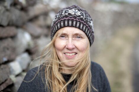 A woman wearing a patterned knit beanie and a dark sweater is smiling outside, with a blurred background of stones and a path. Her blonde hair is partially windblown across her face.