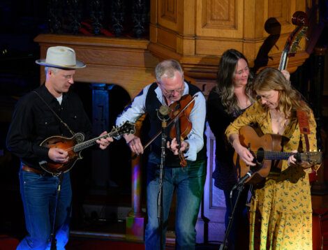 Four musicians play string instruments on stage: a mandolin, a fiddle, a double bass, and a guitar. They are focused on their instruments in a warmly lit wooden setting.