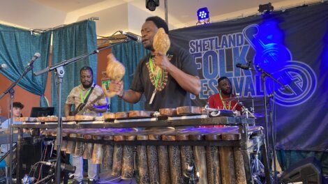 Musicians perform on stage at Shetland Folk Festival. A man in front plays maracas and a xylophone, while others play guitar and drums. A festival banner is visible in the background.