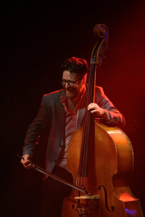 Man playing a double bass with a bow on stage, illuminated by red lighting. He is wearing a suit and glasses and is smiling.