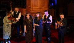 A group of six musicians performs on stage, with members playing a violin, mandolin, and guitar. Two female vocalists sing at the center. The setting appears to be an indoor venue with wooden decor.