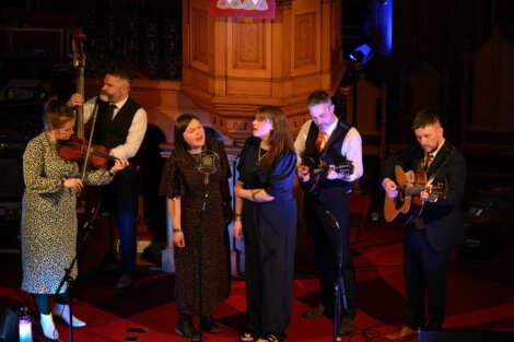 A group of six musicians performs on stage, with members playing a violin, mandolin, and guitar. Two female vocalists sing at the center. The setting appears to be an indoor venue with wooden decor.