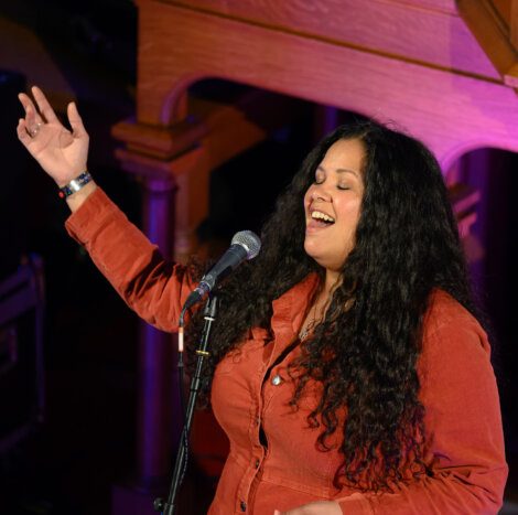 A woman with long curly hair, wearing a red shirt, sings into a microphone with her eyes closed and one arm raised.