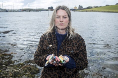 A person stands near a shoreline, holding several collected items in their hands. The background shows water, distant buildings, and rolling hills under a partly cloudy sky.