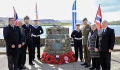 A group of people in formal attire and military uniforms stand in front of a stone memorial with wreaths and flags in the background. The setting is an outdoor location near water.