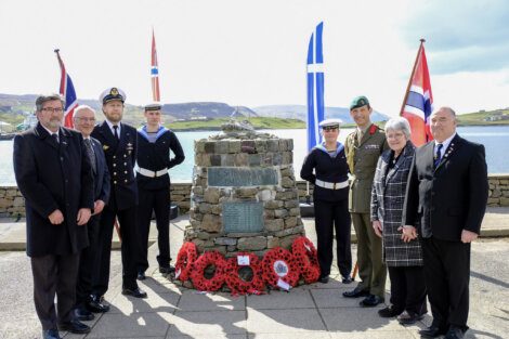 A group of people in formal attire and military uniforms stand in front of a stone memorial with wreaths and flags in the background. The setting is an outdoor location near water.