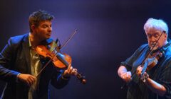 Two men play violins on stage under dramatic lighting. The man on the left wears a blazer, and the man on the right is in a striped shirt.