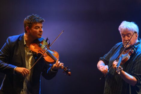 Two men play violins on stage under dramatic lighting. The man on the left wears a blazer, and the man on the right is in a striped shirt.