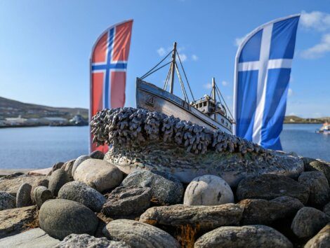 A sculpture of a boat sits on a bed of rocks with Norwegian and Scottish flags in the background under a clear blue sky.