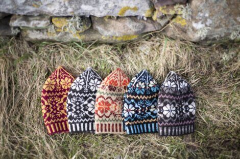 Five knitted hats with colorful patterns are placed on dry grass in front of a stone wall.