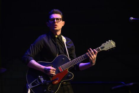 A person wearing glasses and a black shirt is playing a black and red electric guitar on stage.