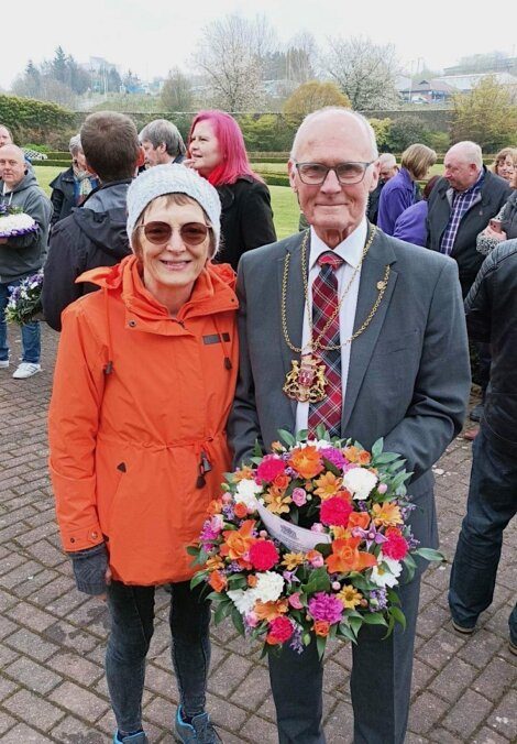 A woman in an orange coat and a man in a suit holding a floral wreath stand together at an outdoor event with people and greenery in the background.